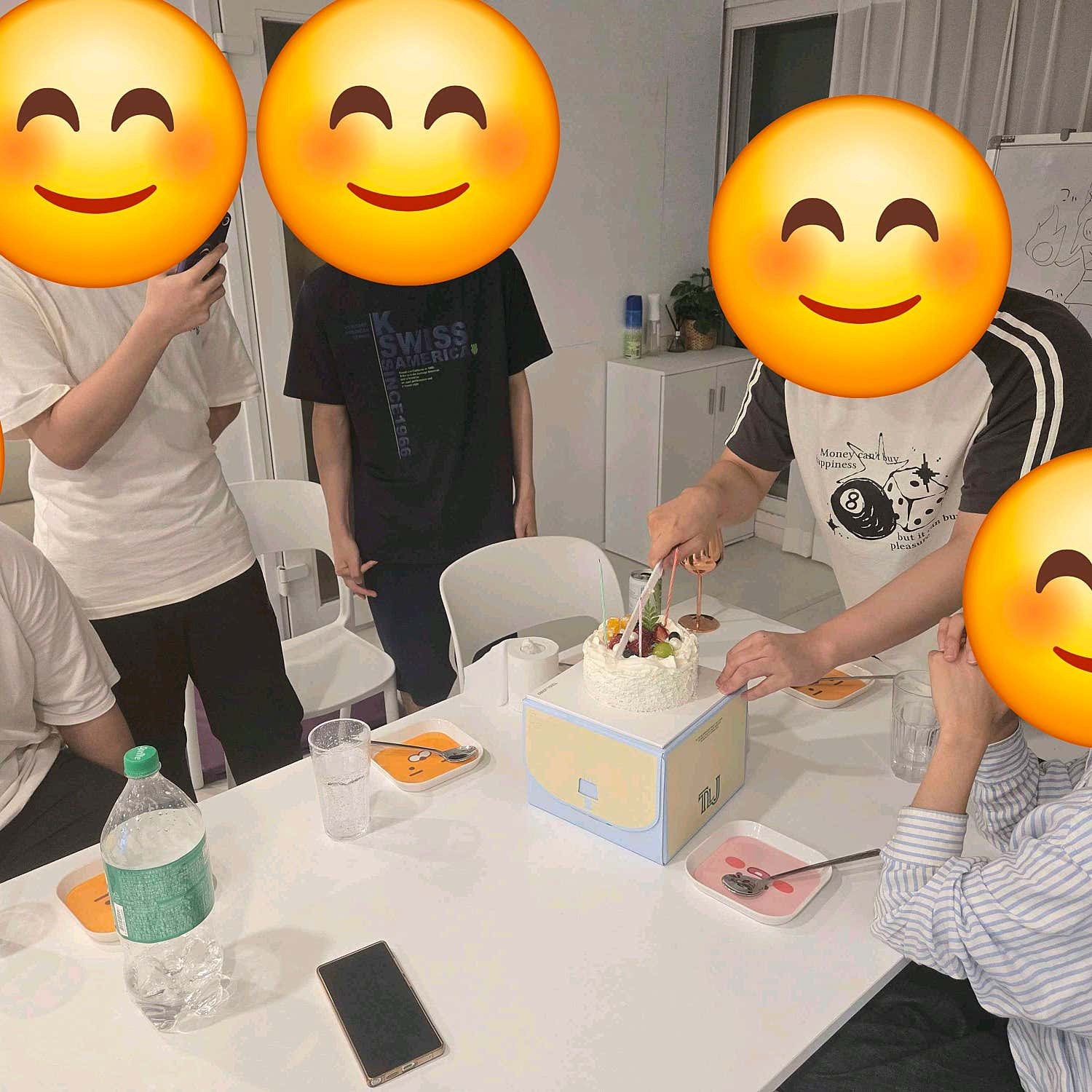 흰 테이블 위 케이크를 자르는 친구들과 음료, 접시, 휴대전화의 꺼진 화면