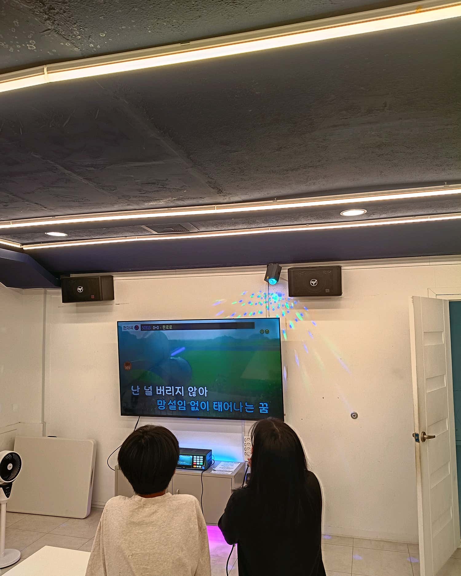 벽면 화면과 노래 기기 앞에 앉아 있는 이용자 두 명의 뒷모습