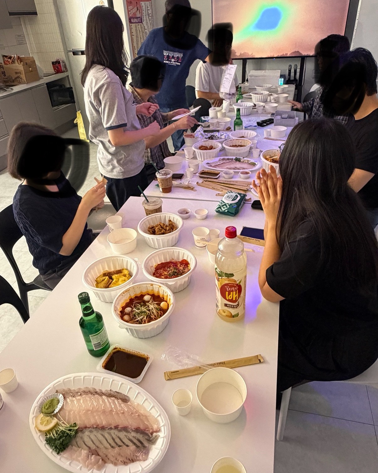 긴 테이블 위 여러 포장 음식과 화면 앞에 모인 단체